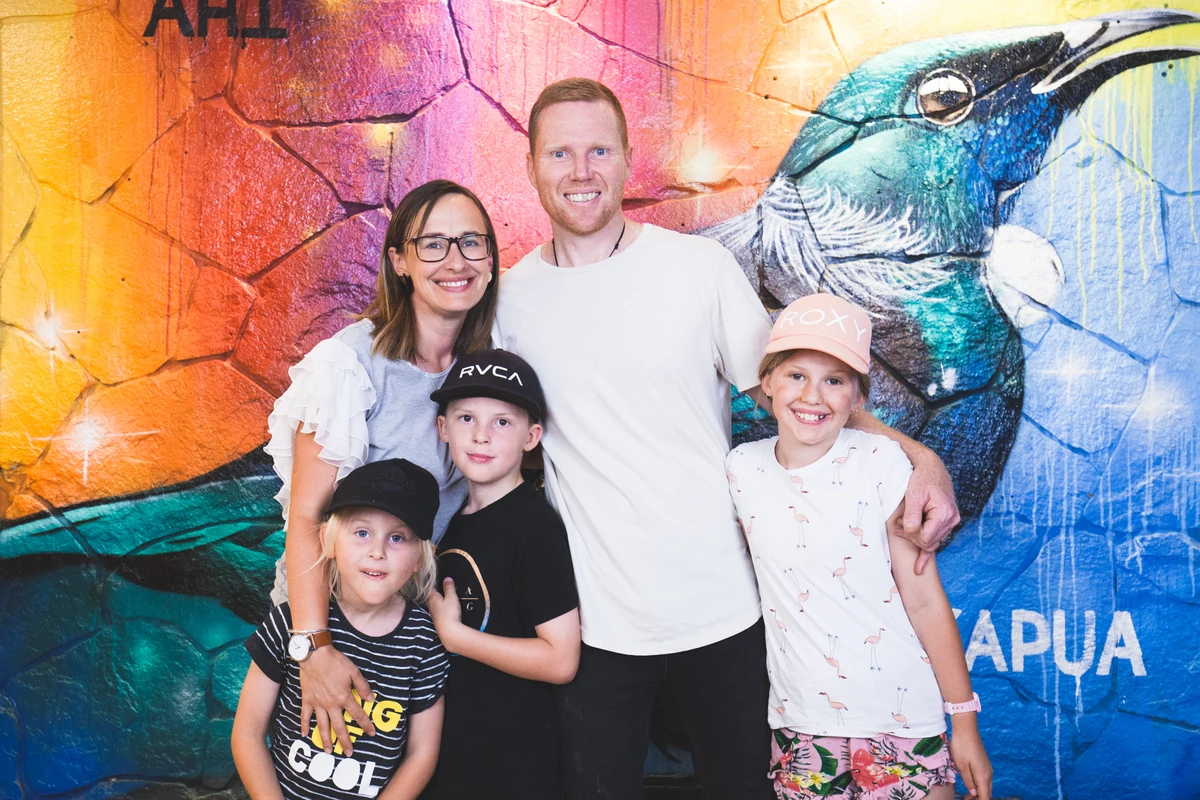 A family of five smiling together in front of a colourful bird mural.