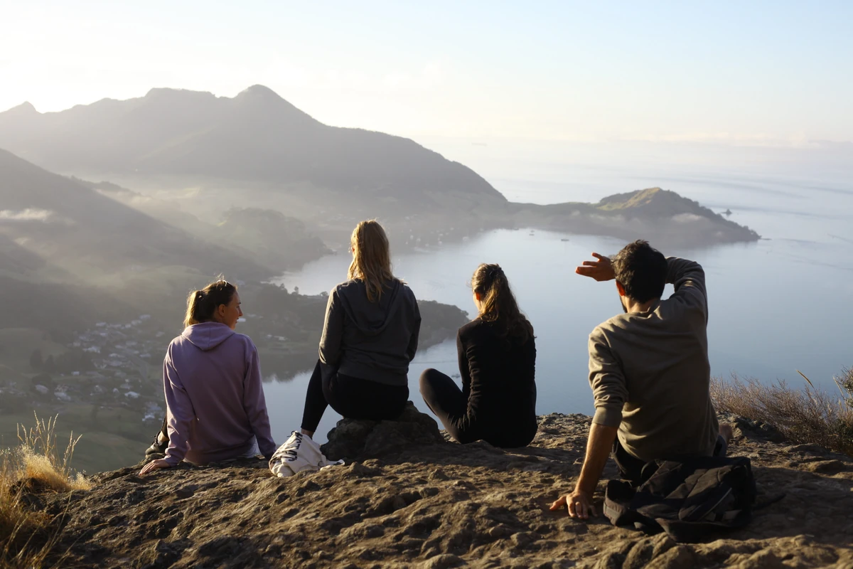 Four people looking out from a rocky hilltop over a harbour and coastal hills.