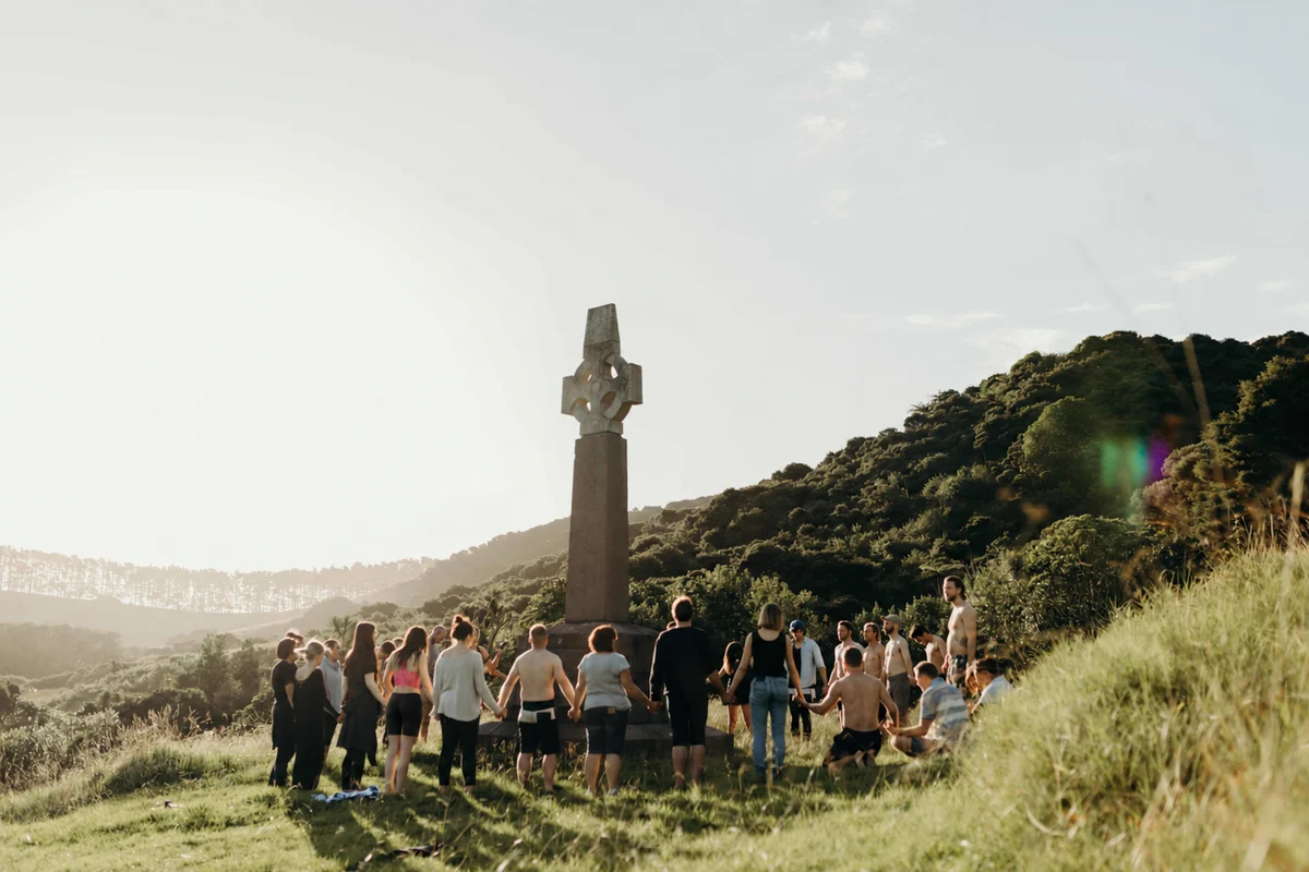 People holding hands in a circle beside Marsden Cross in sunlit coastal hills.